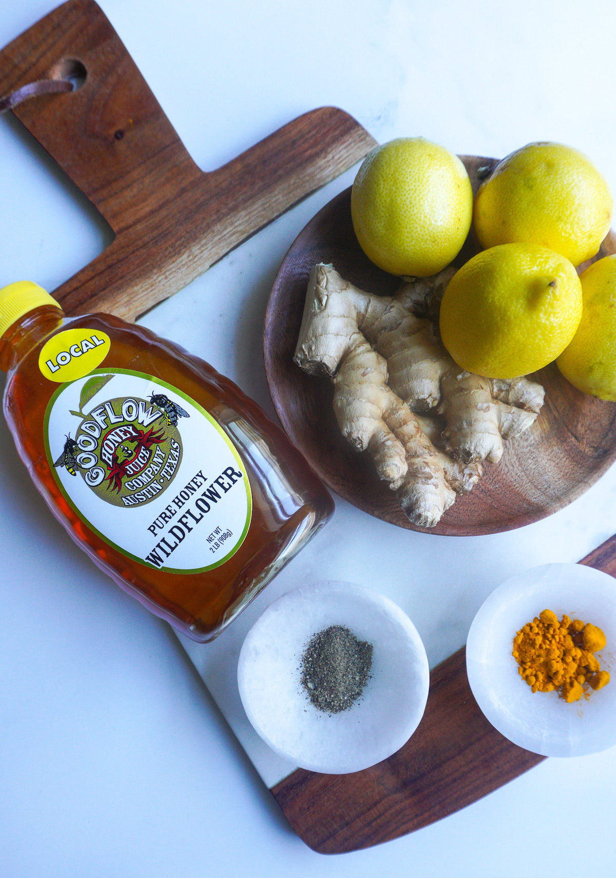 A bottle of honey, some lemons, a knob of ginger, turmeric and pepper on a cutting board