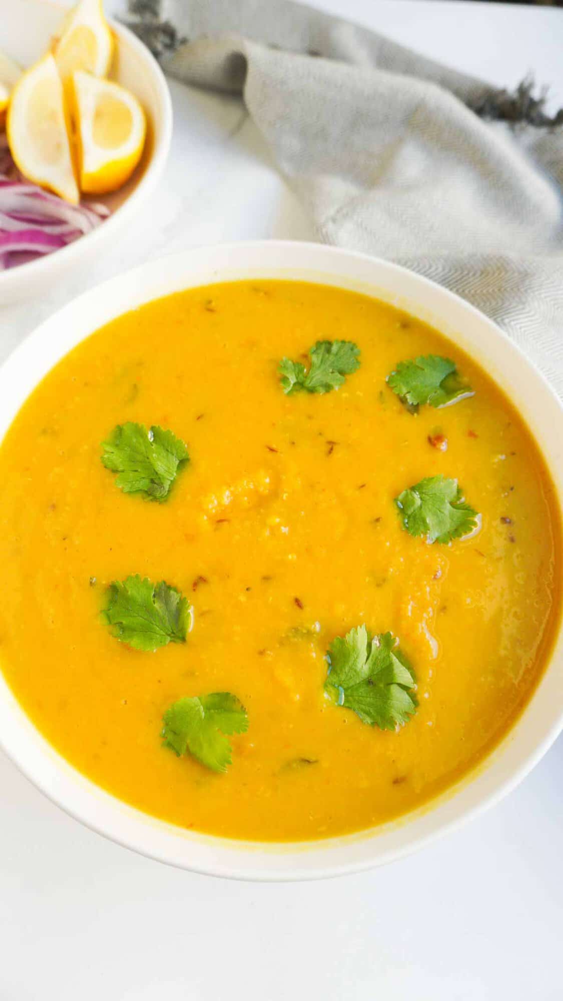 A bowl of tadka dal with cilantro garnish. A napkin and a bowl of lemon wedges and red onion off to the side.