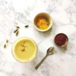 a glass of turmeric milk with some cardamoms, saffron and turmeric on the side in condiment dishes