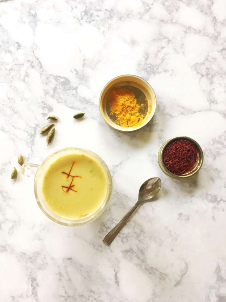 a glass of turmeric milk with some cardamoms, saffron and turmeric on the side in condiment dishes