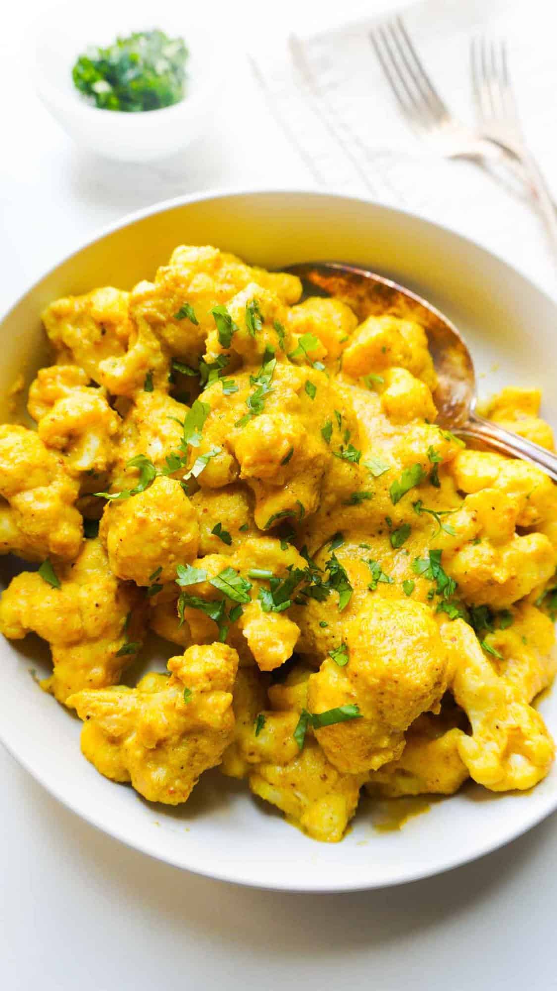 cauliflower cashew curry in a serving bowl with cilantro garnish bowl on the side and a couple of forks on a napkin to the right