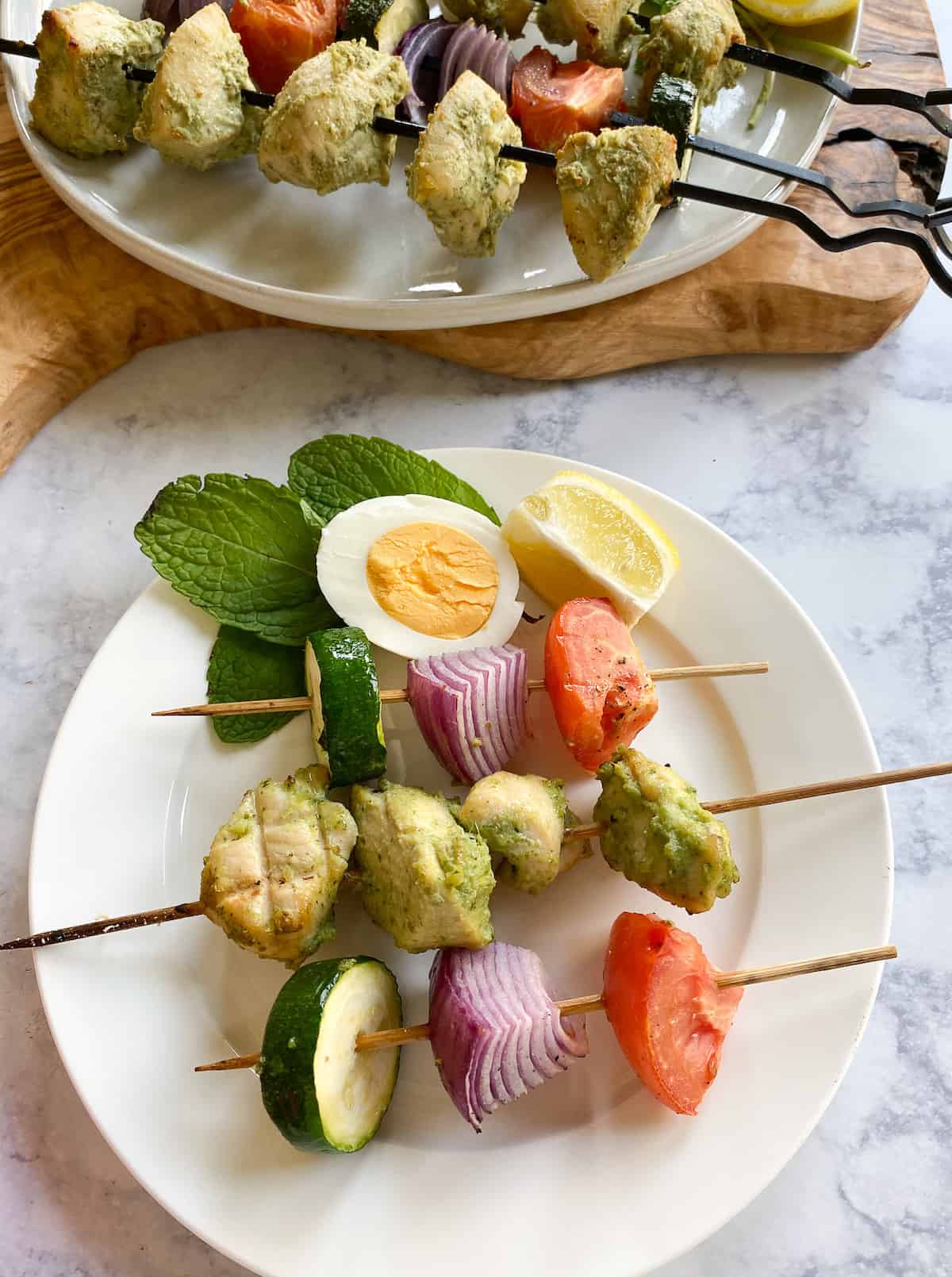 Skewers of zucchini, tomatoes and onions and hara masala chicken skewers on a plate with a boiled egg and lemon wedge with a few mint leaves as garnish