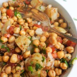 A bowl of chickpeas and potatoes coated in imli/tamarind chutney and garnished with tomatoes, green chili, cilantro.