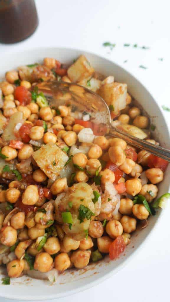A bowl of chickpeas and potatoes coated in imli/tamarind chutney and garnished with tomatoes, green chili, cilantro.