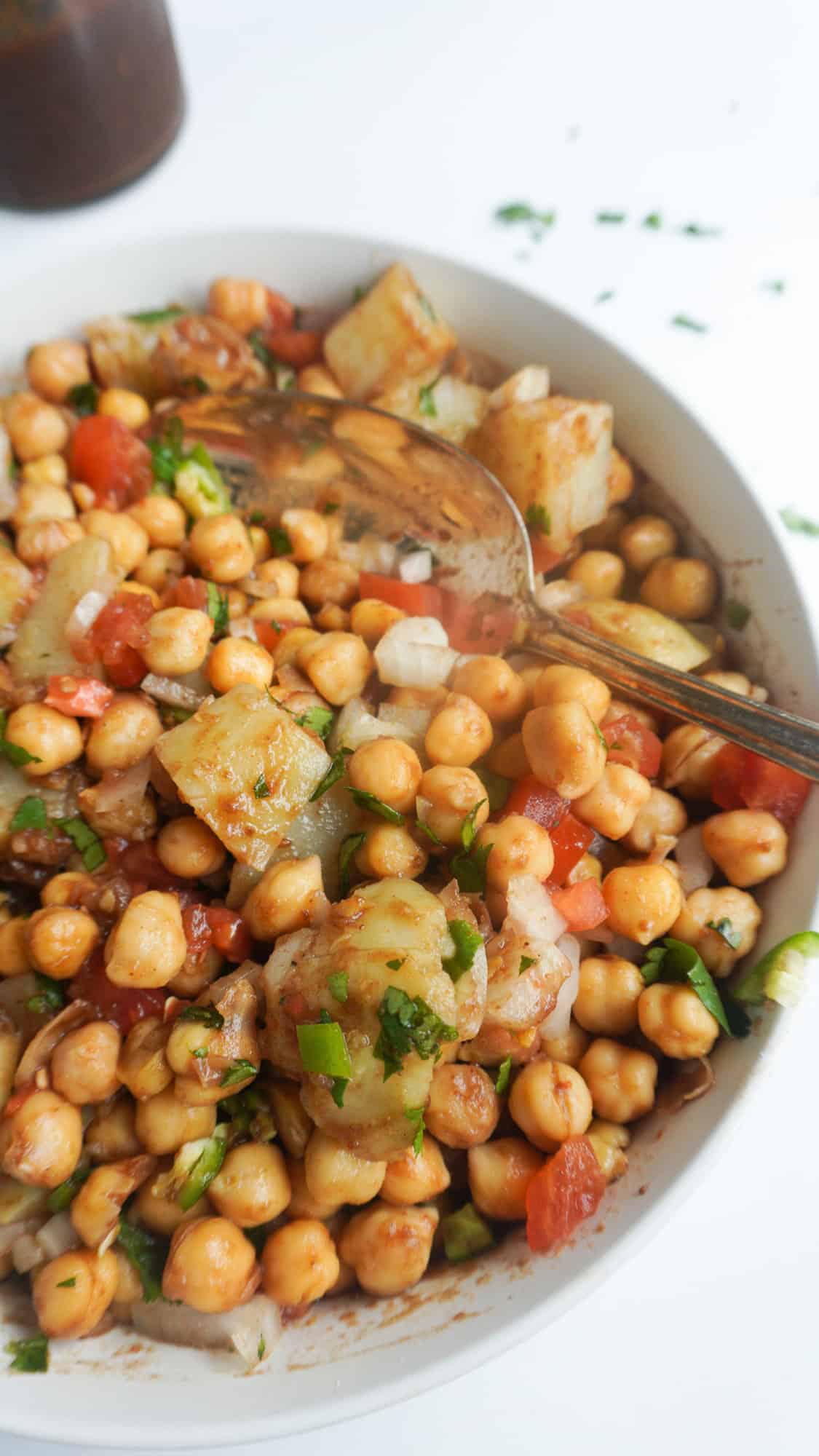 A bowl of chickpeas and potatoes coated in imli/tamarind chutney and garnished with tomatoes, green chili, cilantro. 