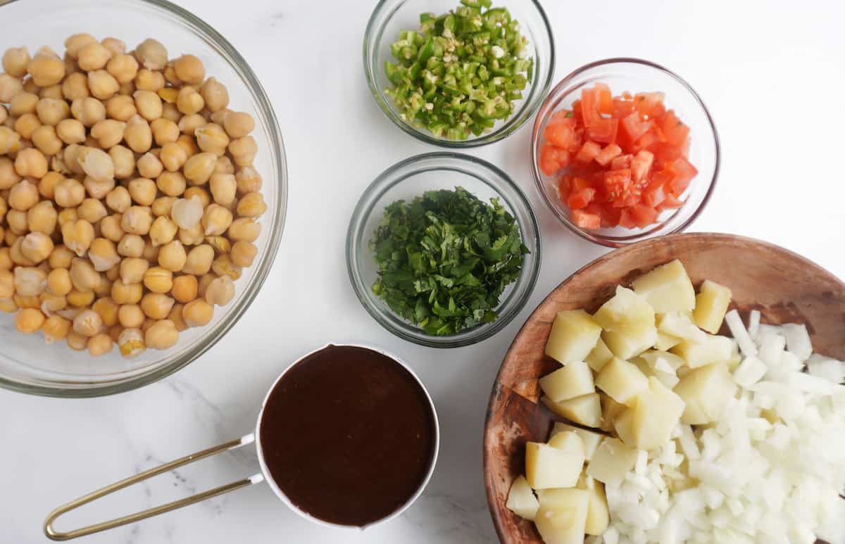 A bowl of chickpeas, a bowl of boiled potato, diced onion and a measuring cup with tamarind/imli chutney also pictured, smaller bowls of cilantro, tomatoes and green chili pepper.