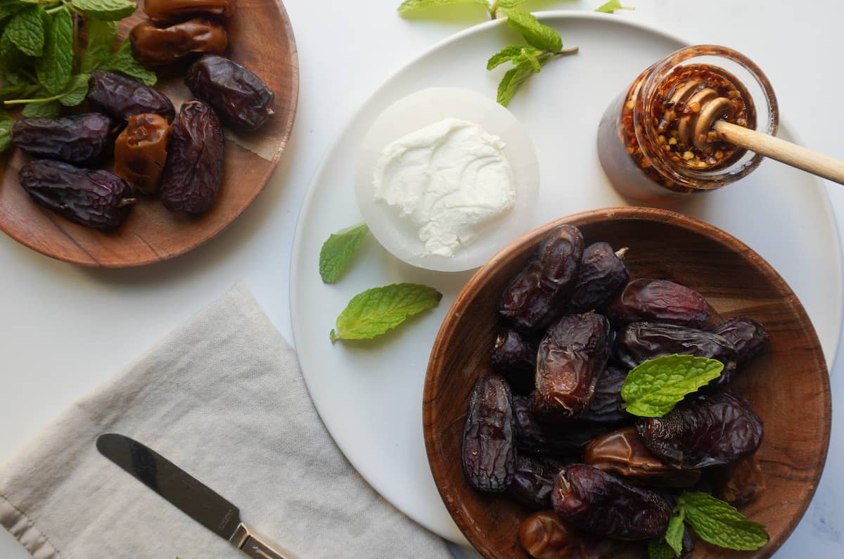 A plate with dates in a wooden bowl, goat cheese in a bowl, a jar of honey and a honey spoon and mint leaves