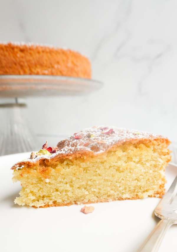 A slice of cardamom cake on a plate with a cake on a pedestal platter in the background