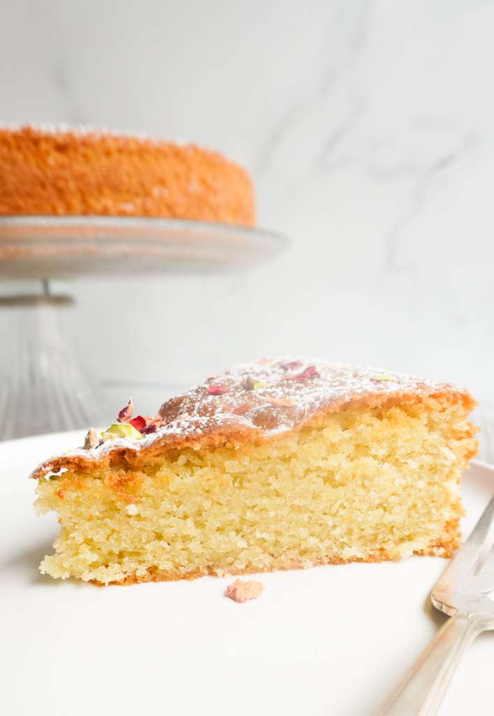 A slice of cardamom cake on a plate with a cake on a pedestal platter in the background