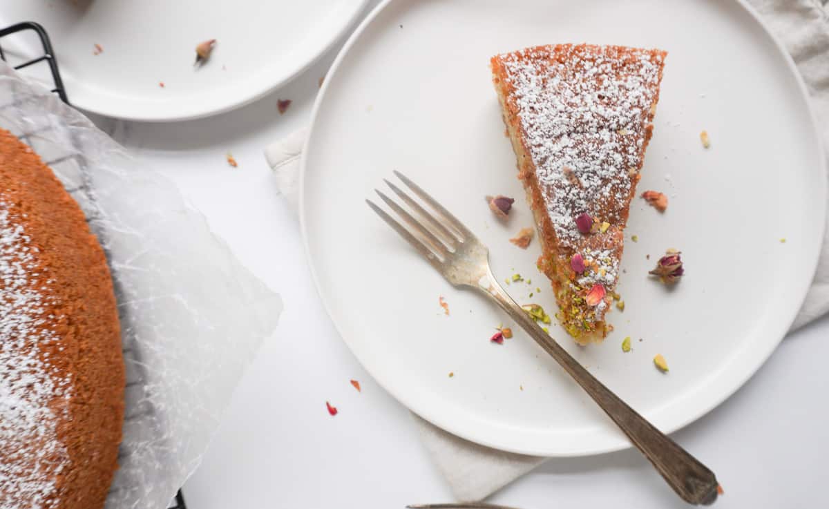 A plate with a fork and a slice of cardamom cake and a wire rack with partial view of the rest of the cake. There is another plate to the side.