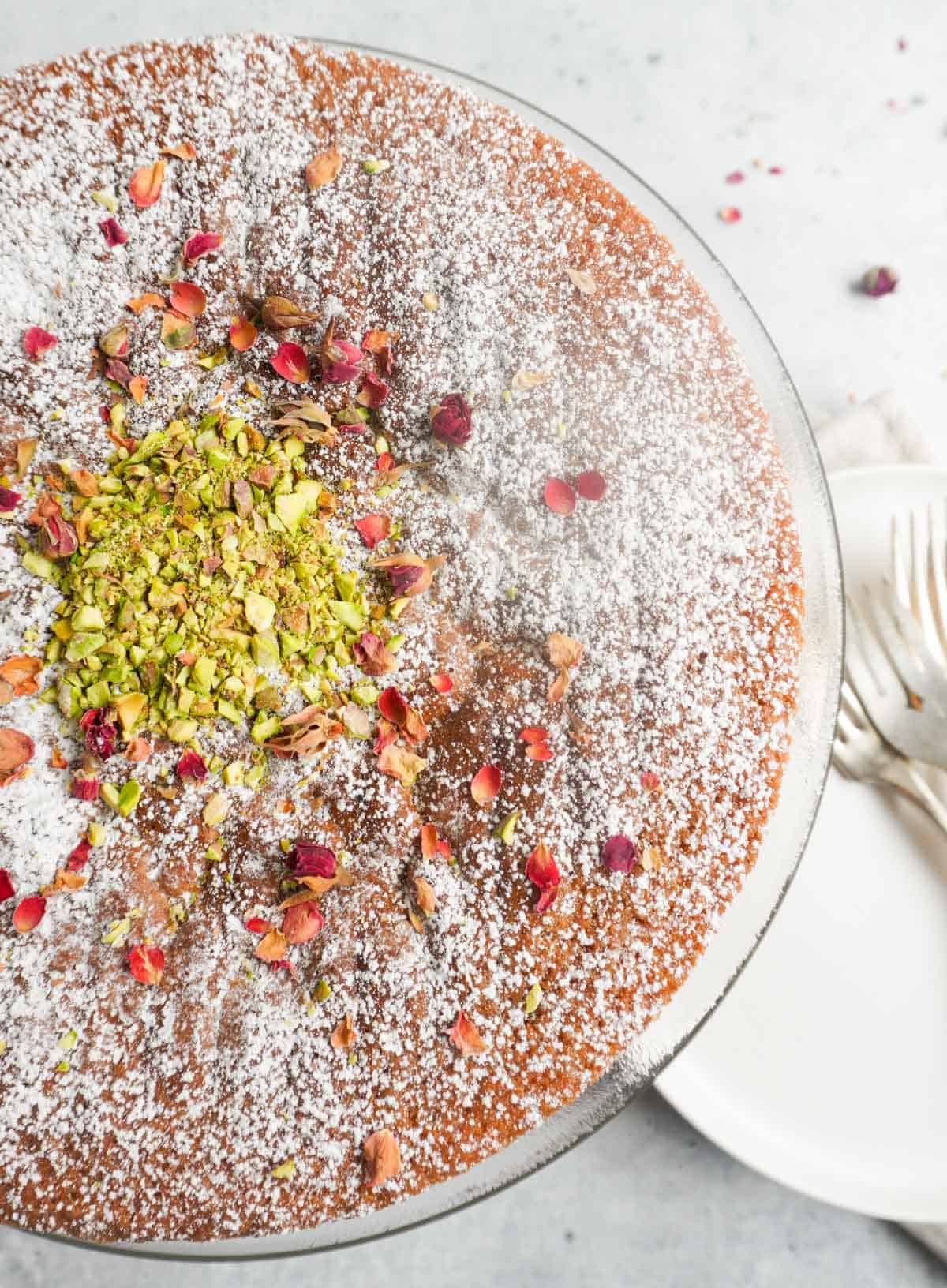 Gluten free cardamom cake on platter with chopped pistachio garnish in center and rose petals with a plate and forks to the side