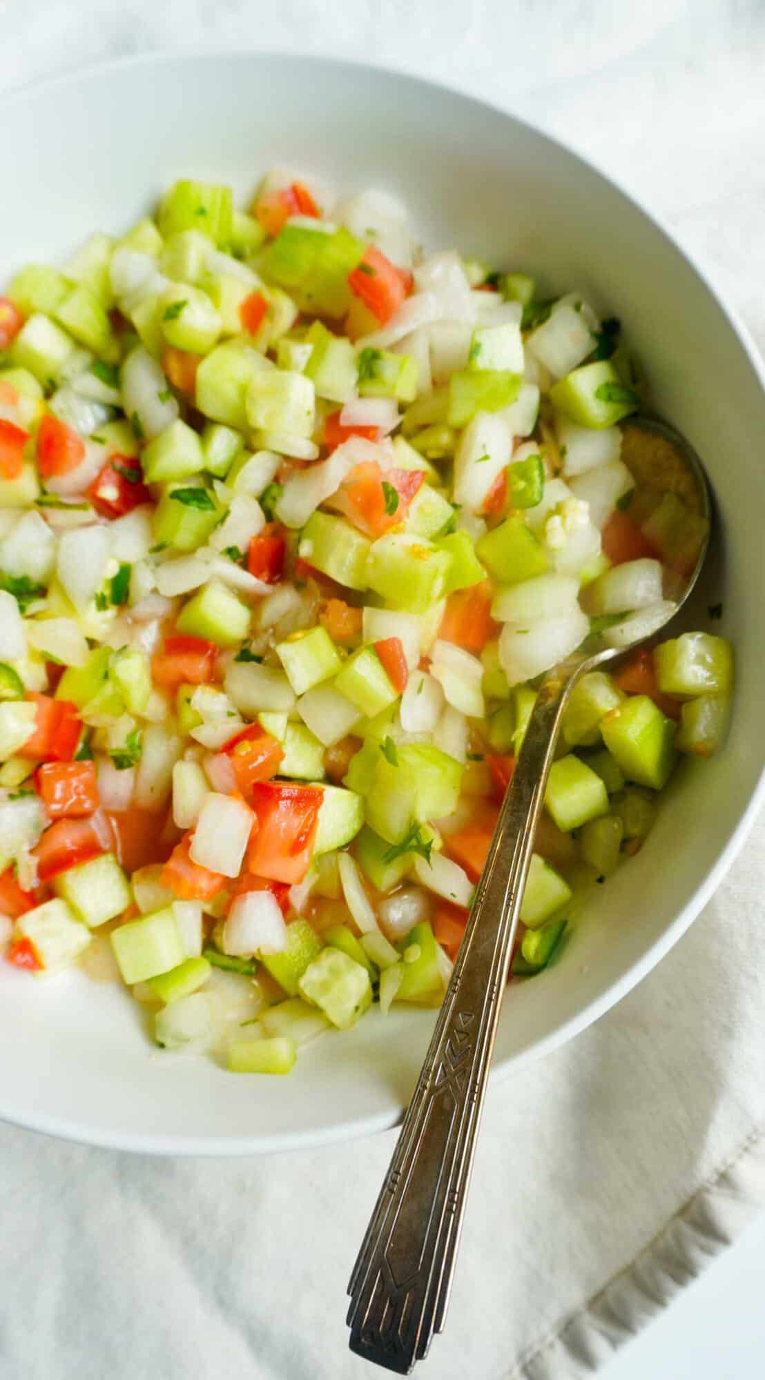 A bowl of kachumbar - finely diced cucumbers, tomatoes and onions with a spoon.