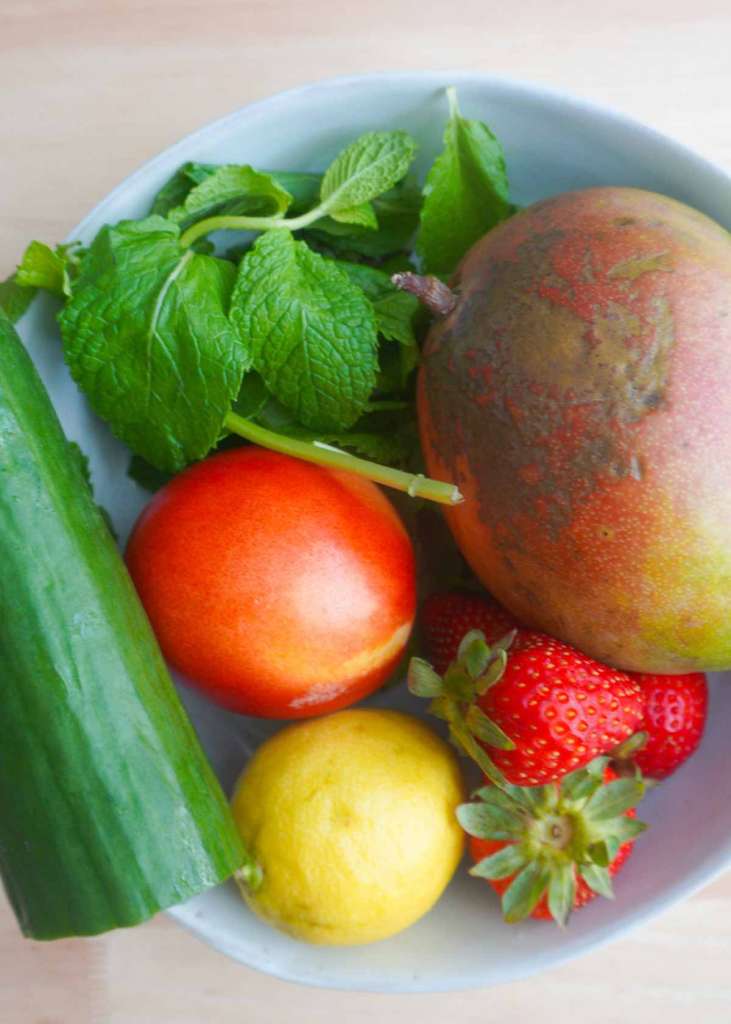 Cucumber, mint sprig, nectarine, lemon, strawberries and mango in a bowl.