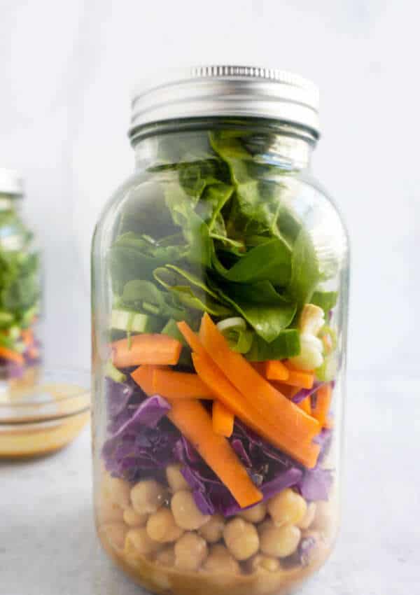 A large glass mason jar with dressing, vegetables (carrot, spinach, red cabbage, spinach and chickpeas) layered. Another jar in the background as well as dipping bowl with peanut dressing
