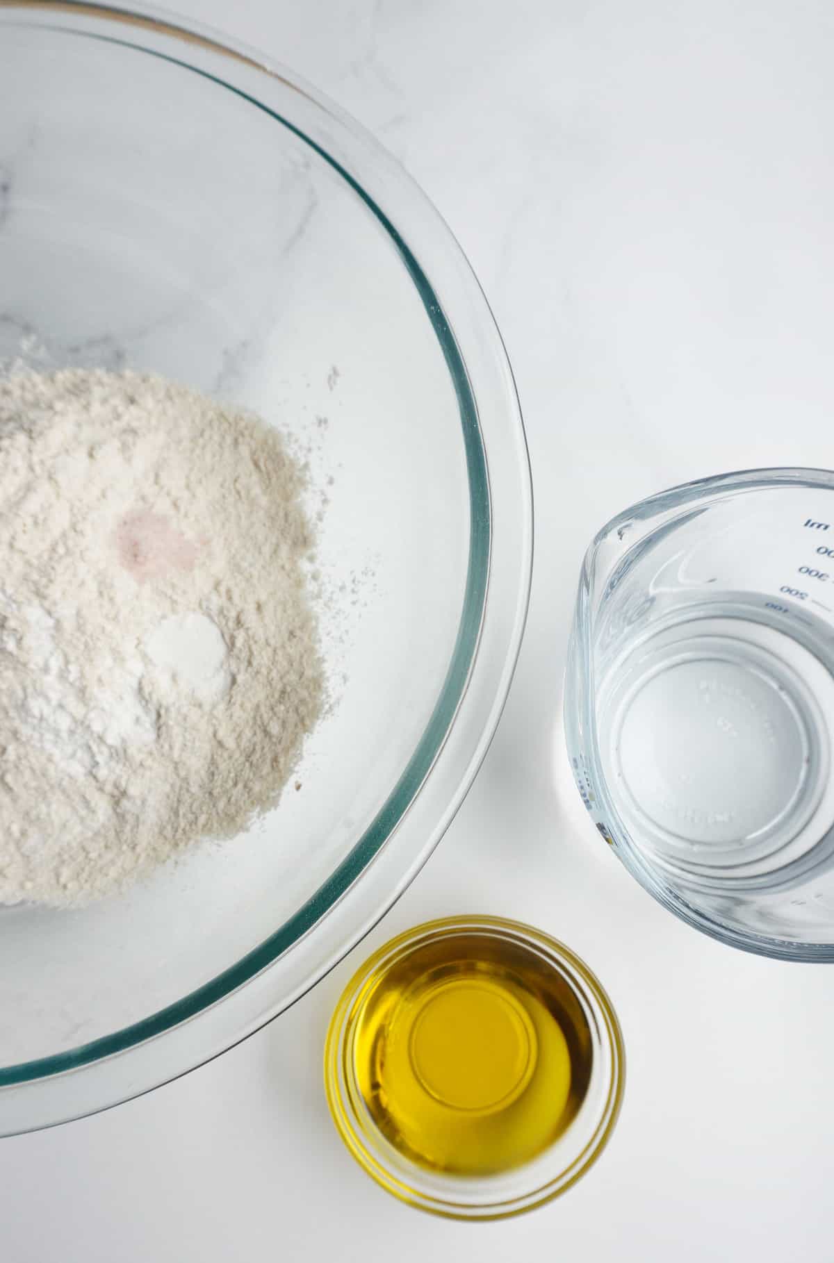 a large glass bowl with barley flour, salt and baking soda and a measuring cup filled with water and a dipping bowl filled with olive oil 