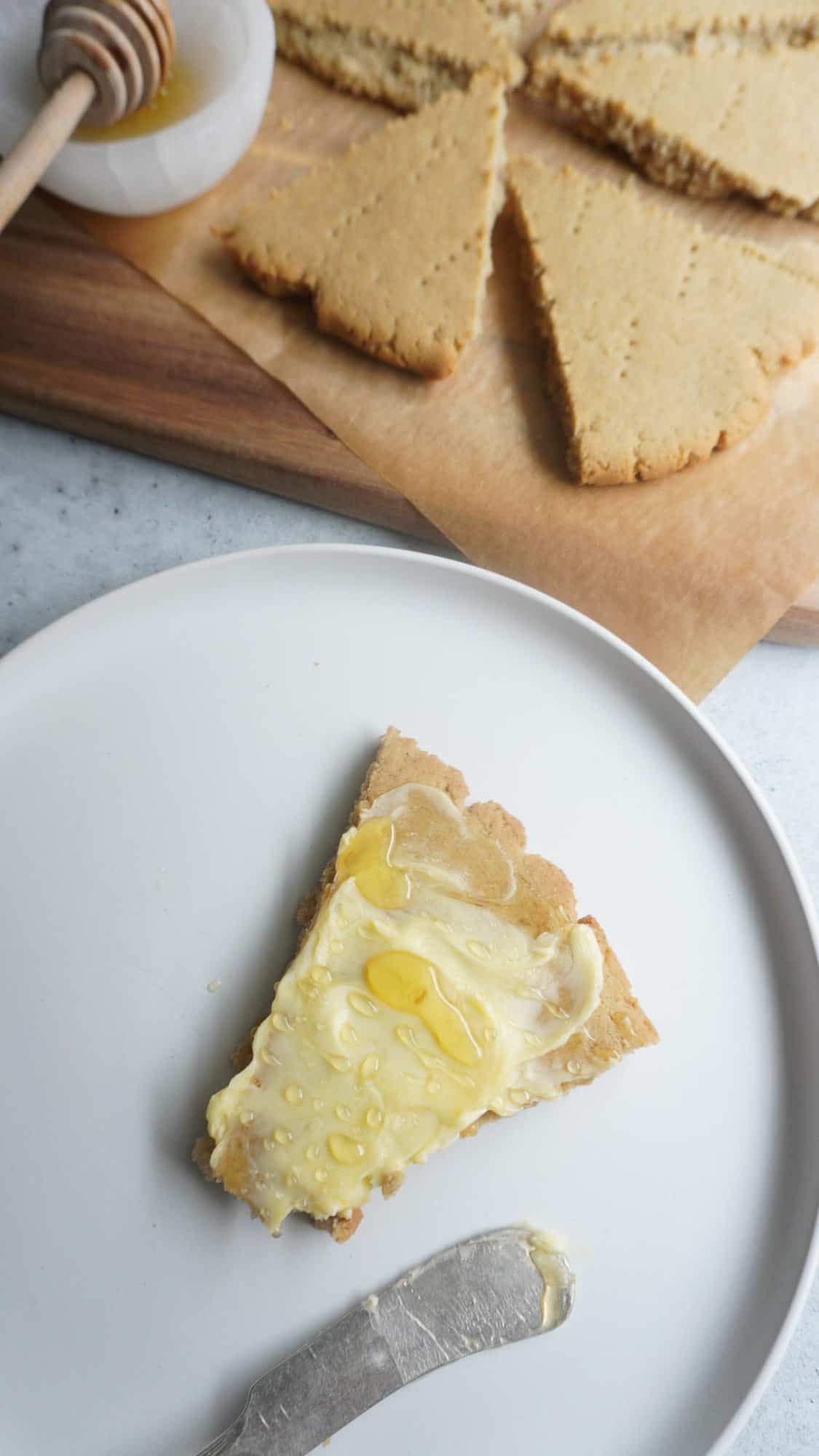 A plate with a piece of barley bread covered with butter and honey. A chopping board with more pieces of bread to the side 