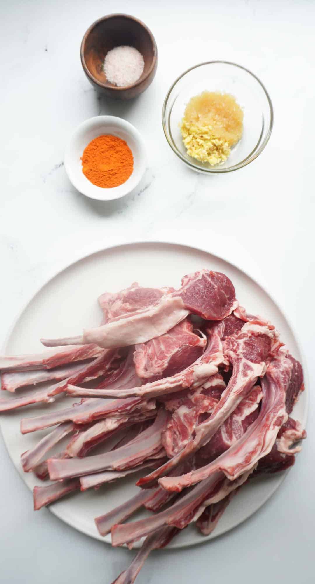 Ingredients for masala chops. A plate full of uncooked lamb chops with bowls of ginger, garlic, pink salt and red chili powder.