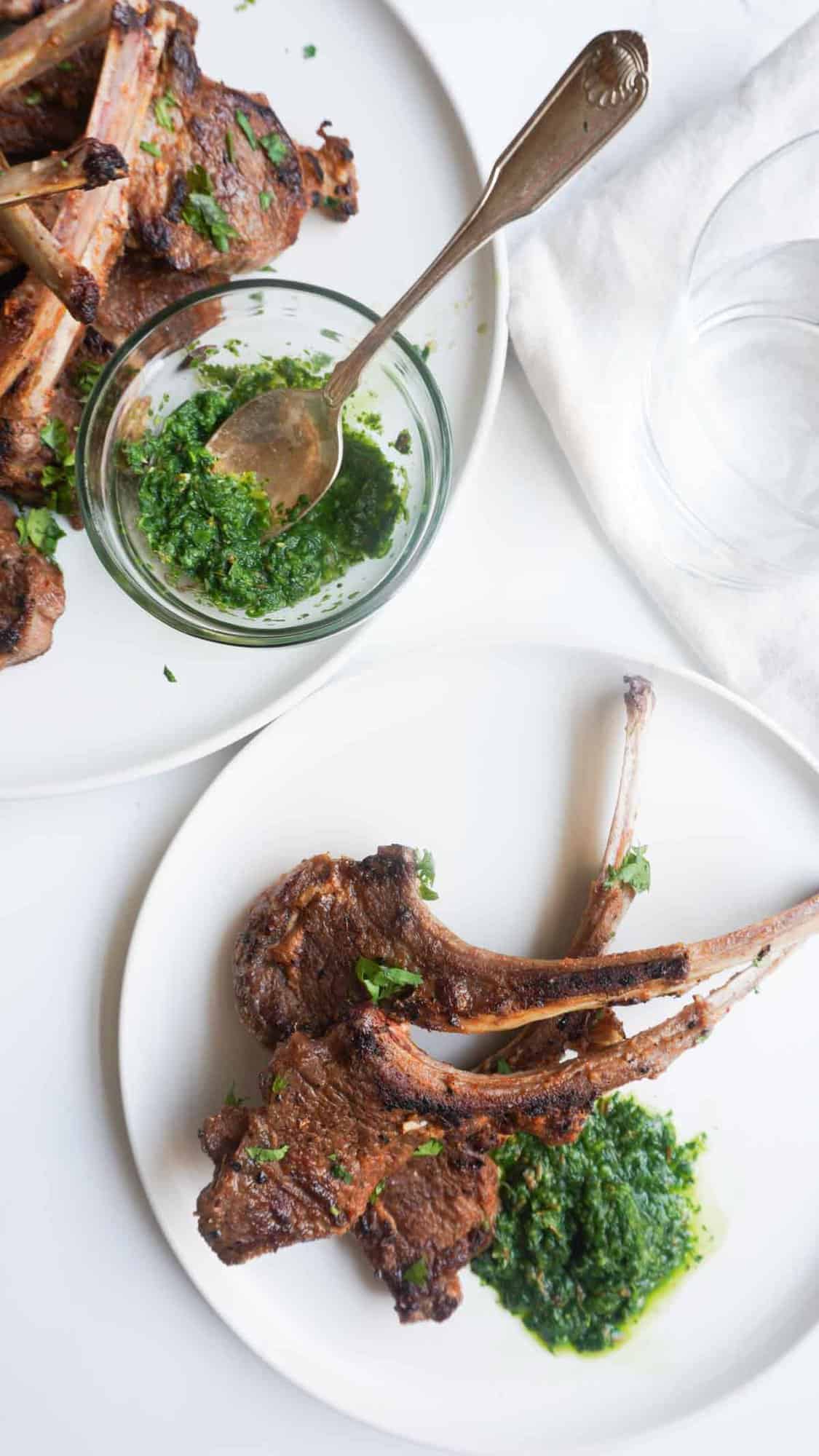 A plate with three lamb chops and some green mint chutney. A platter with lamb chops and condiment bowl with chutney and a small spoon.