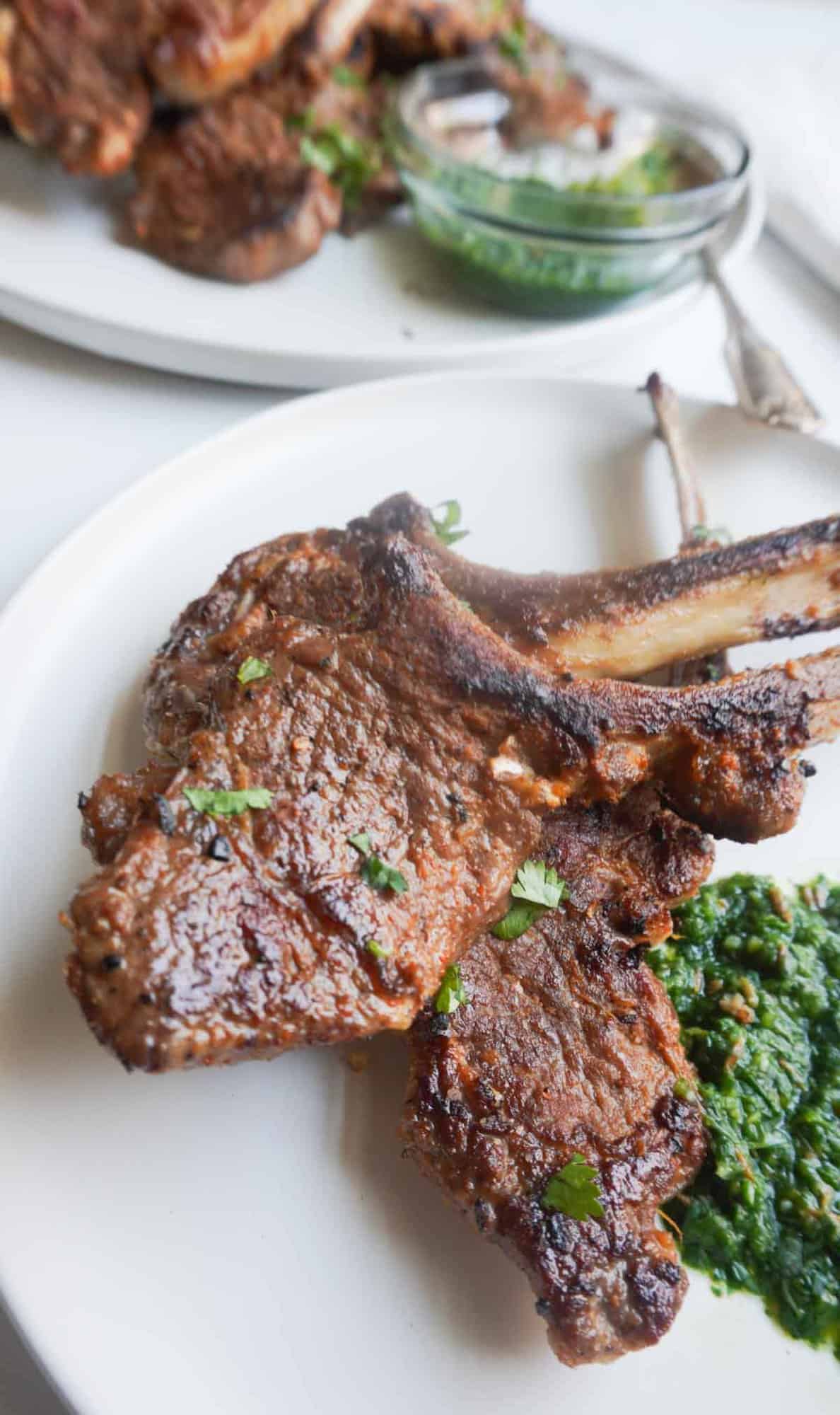 a plate of 3 lamb chops with mint chutney. a platter of lamb chops and a dipping bowl of mint chtuney and a spoon in the background