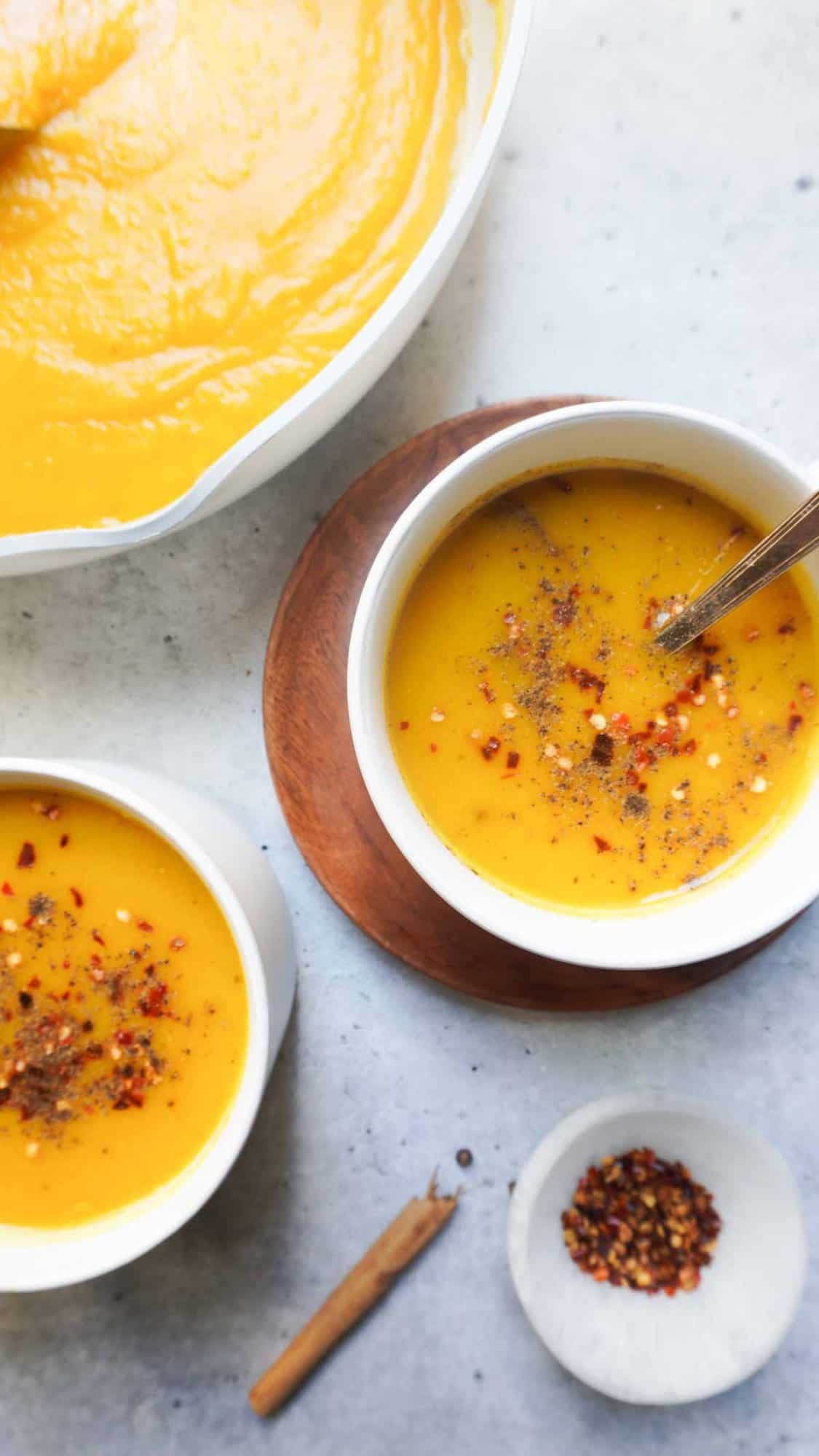 2 bowls of pumpkin soup with a pot of soup in the left far corner and spices in front