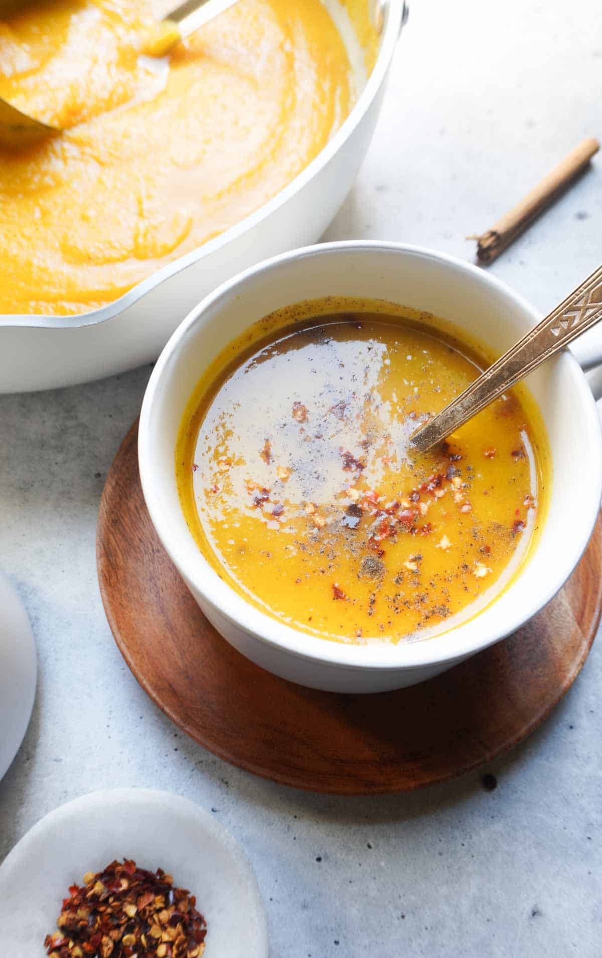 a cup of AIP butternut squash soup with a pot in the back ground and red pepper flakes dipping bowl in front left corner
