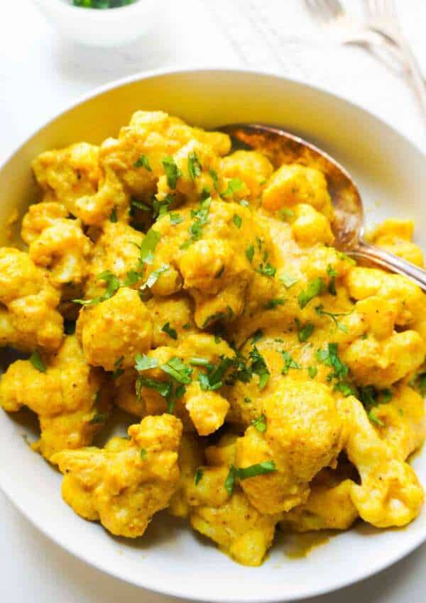 A bowl of cashew cauliflower curry with some forks, a napkin and cilantro garnish on the side.