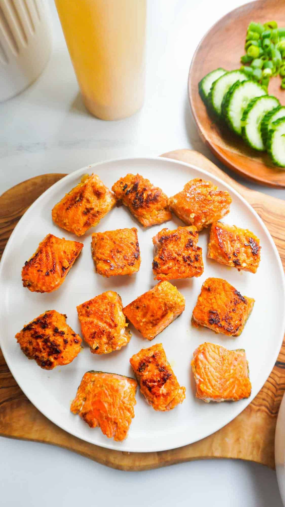 fA plate of salmon tikka bites with spicy mayo squirt bottle above it and a plate of extra cucumbers and green nions on the side.