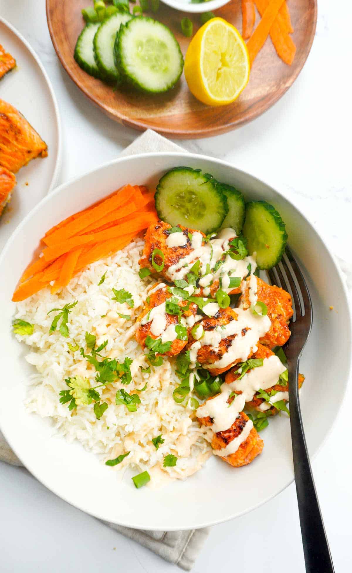 Spicy salmon bowl. Cucumbers, half a lemon and carrot ribbons on a wooden plate to the side.