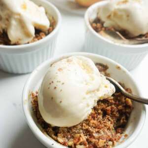 three individual serving bowls of apple crisp with vanilla ice cream and some apples in the background