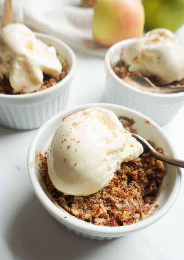 three individual serving bowls of apple crisp with vanilla ice cream and some apples in the background