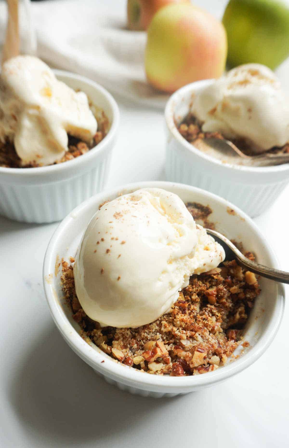 three individual serving bowls of apple crisp with vanilla ice cream and some apples in the background