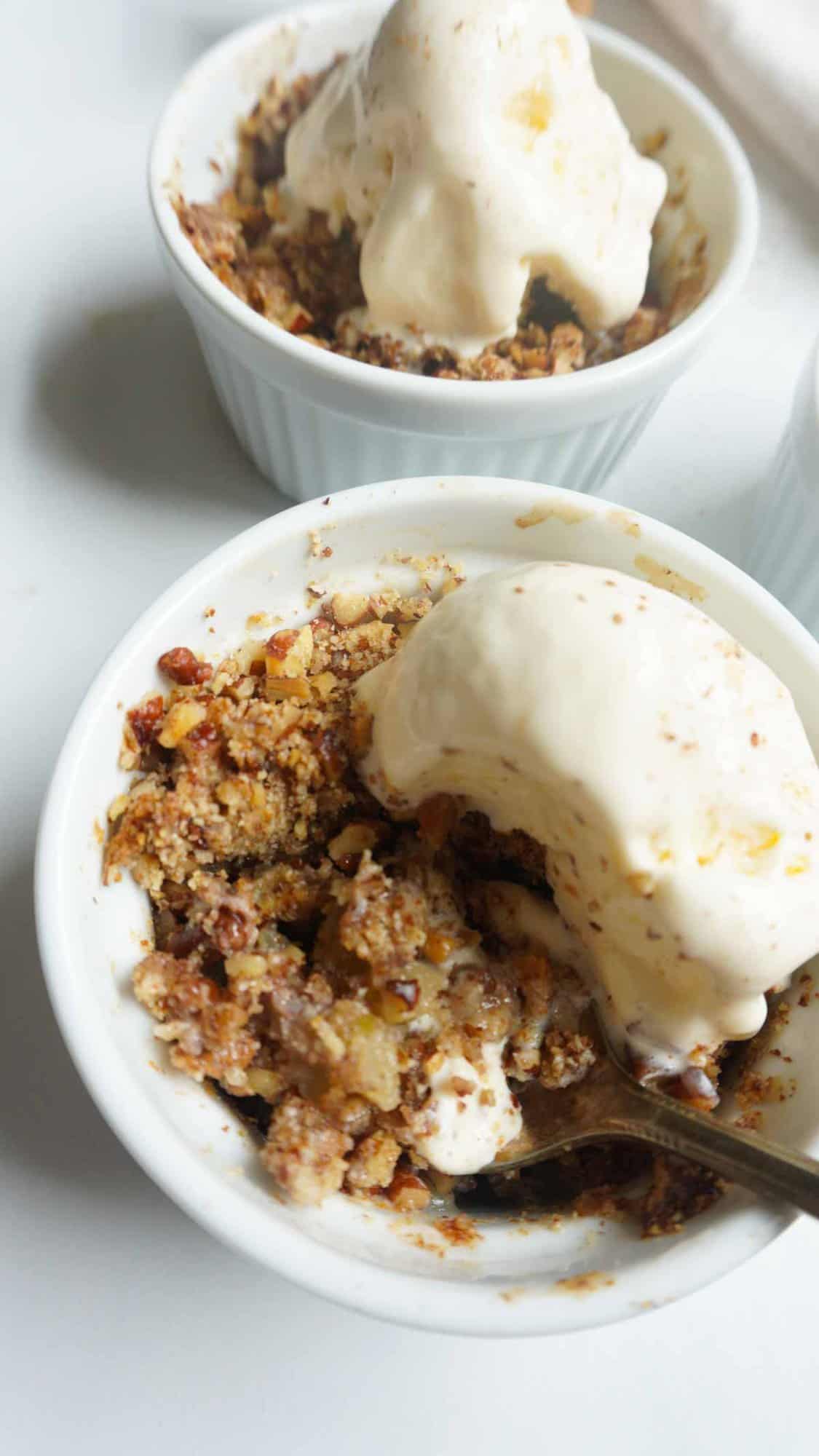 A ramekin with apple crisp and a spoon and a scoop of vanilla ice cream. Another bowl of apple crisp in back ground