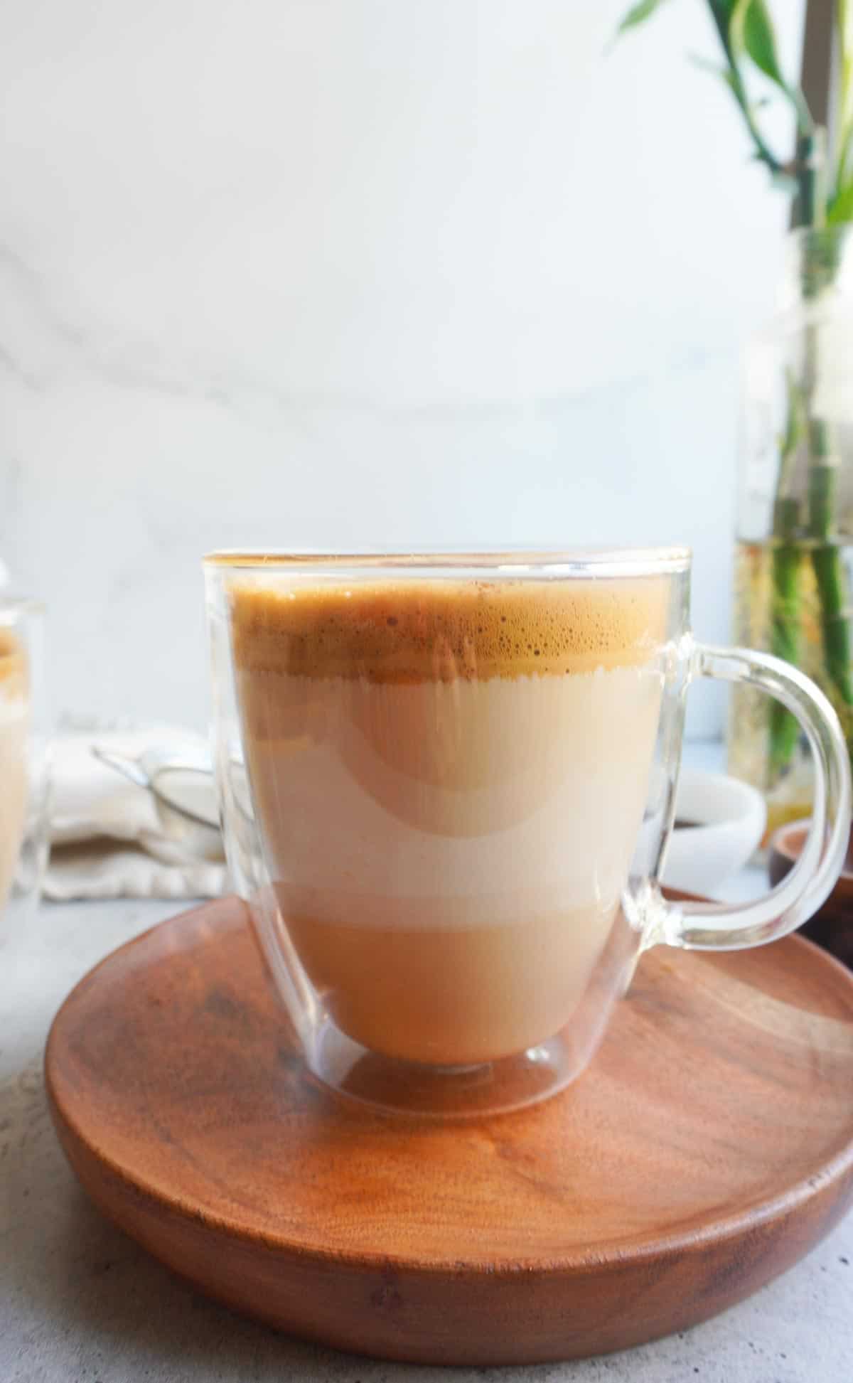 A clear mug with desi coffee on a wooden saucer.