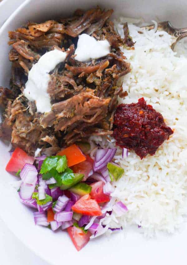 Close up shot of lamb bowl with white rice, cucumber tomato salad, garlic dressing and harissa