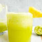 A close up of pineapple cucumber smoothie with pineapple chunk garnish. More smoothie in a jug and pineapple chunks in the background