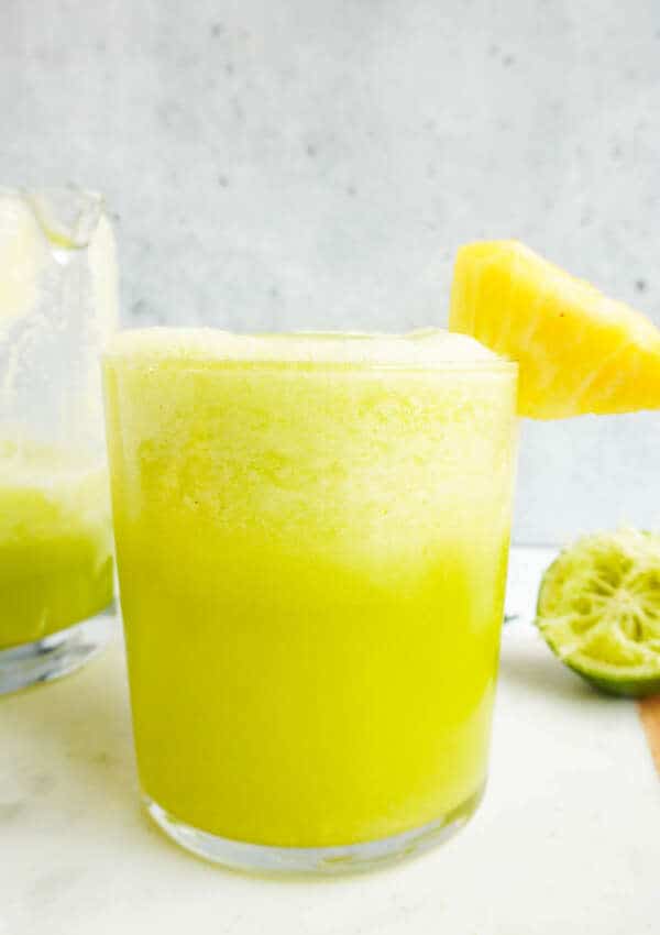 A close up of pineapple cucumber smoothie with pineapple chunk garnish. More smoothie in a jug and pineapple chunks in the background