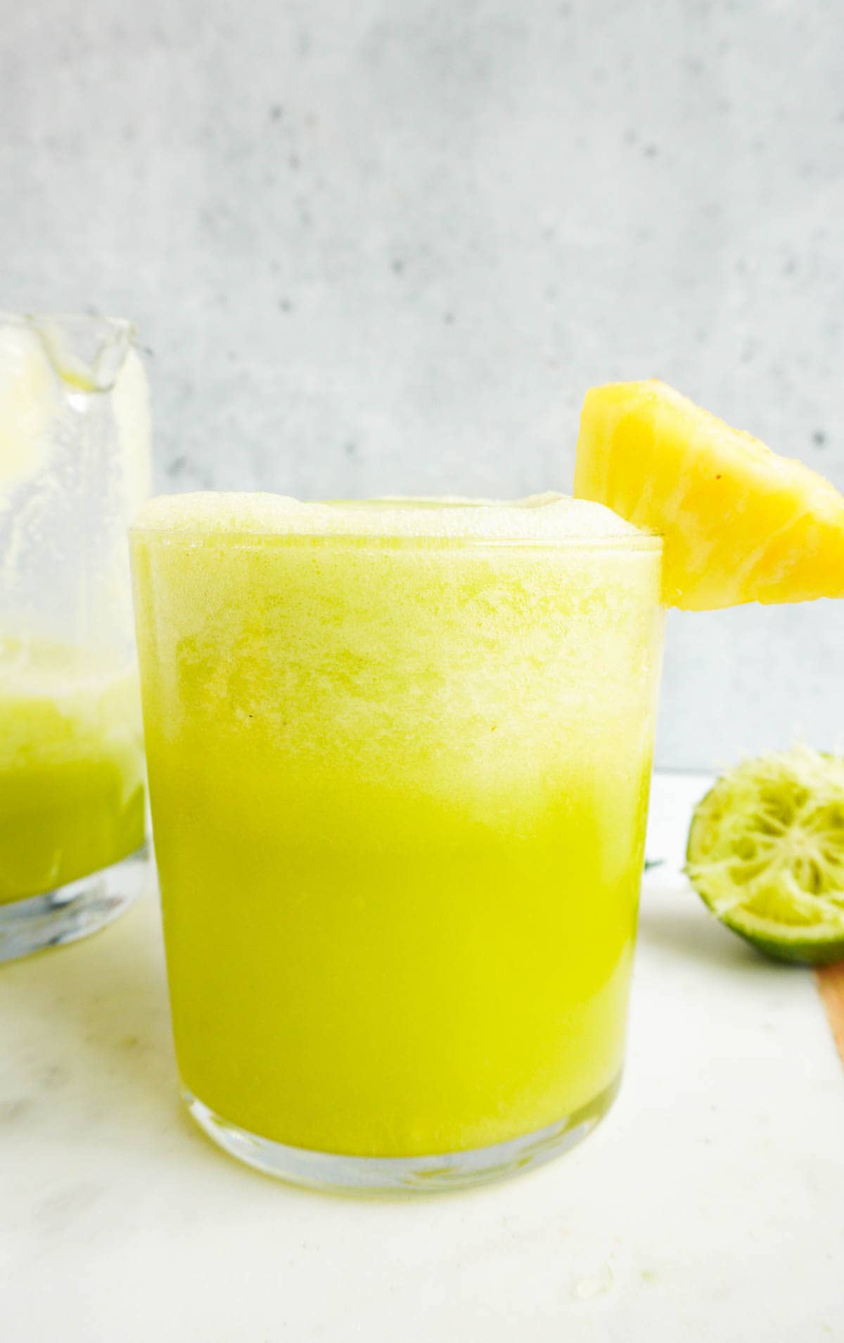 A close up of pineapple cucumber smoothie with pineapple chunk garnish. More smoothie in a jug and pineapple chunks in the background