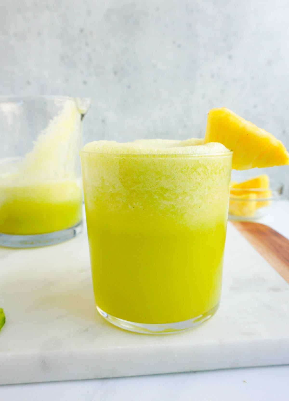 A glass of pineapple cucumber smoothie with a pineapple chunk garnish. A jug of smoothie on the side with a bowl of pineapple chunks on the right.