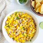 A bowl of mango corn salsa, a bowl of chips a half lime and cilantro
