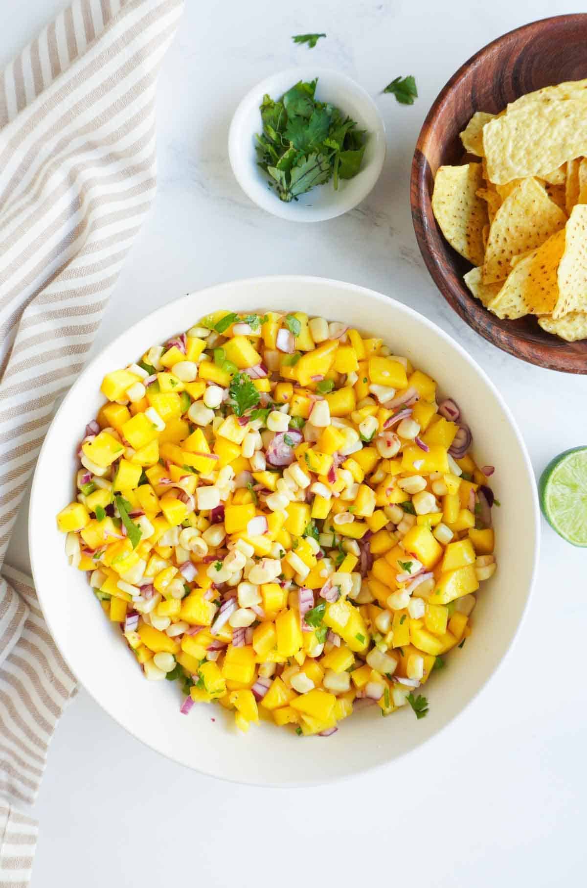 A bowl of mango corn salsa, a bowl of chips a half lime and cilantro