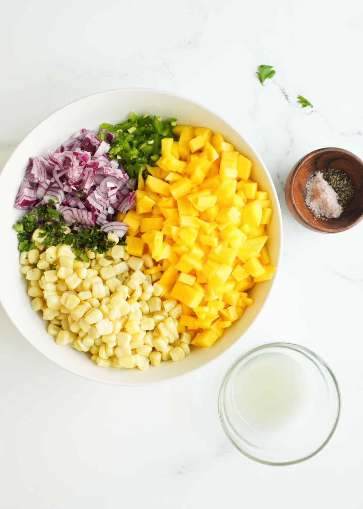Ingredients for mango corn salsa in a bowl and seasonings in dipping bowls to the side.