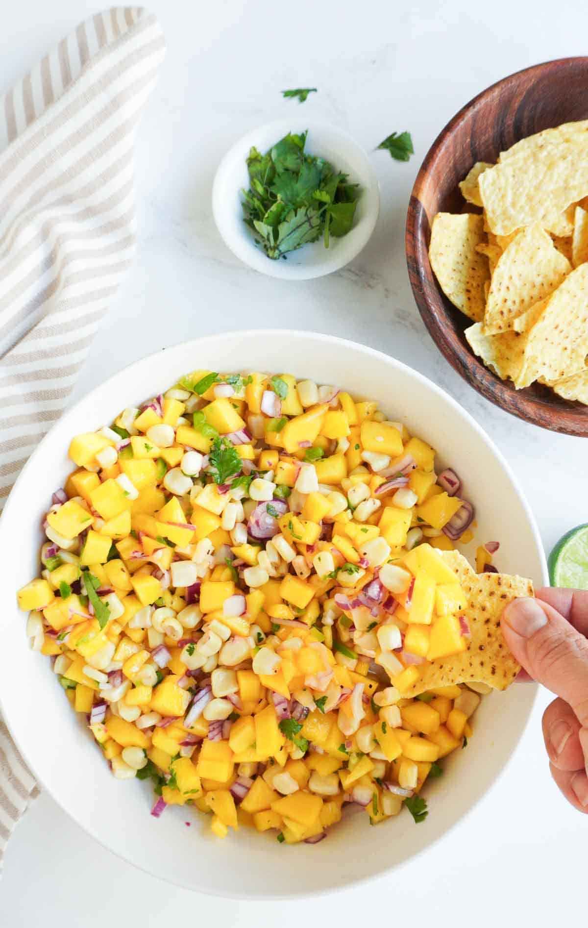 A bowl of mango corn salsa with a hand holding a chip and some salsa above it.