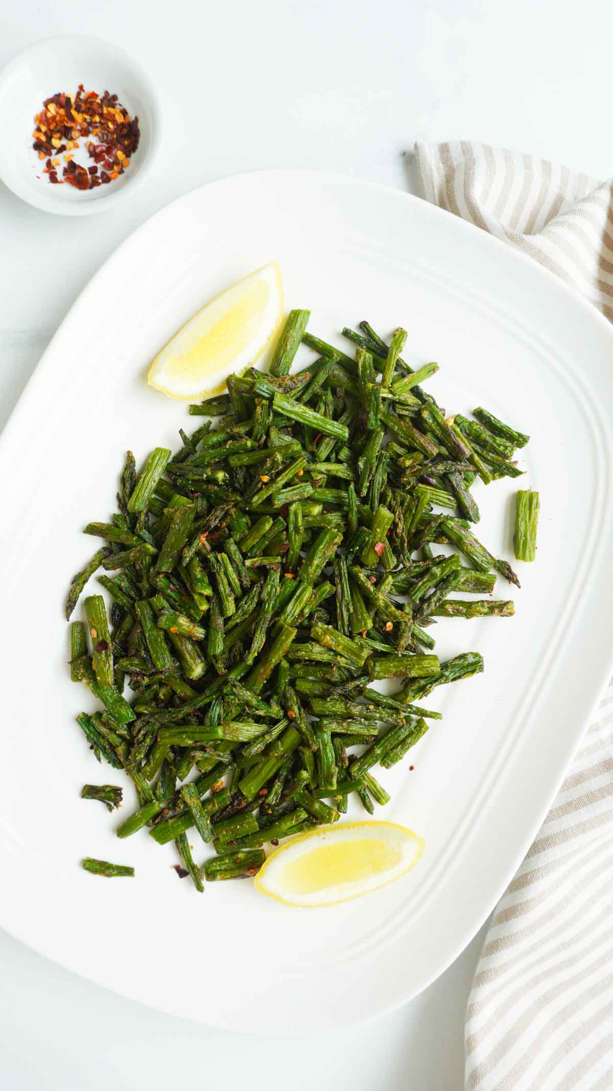 air fried asparagus in a platter with some lemon and crushed red pepper flakes in a bowl on the side.