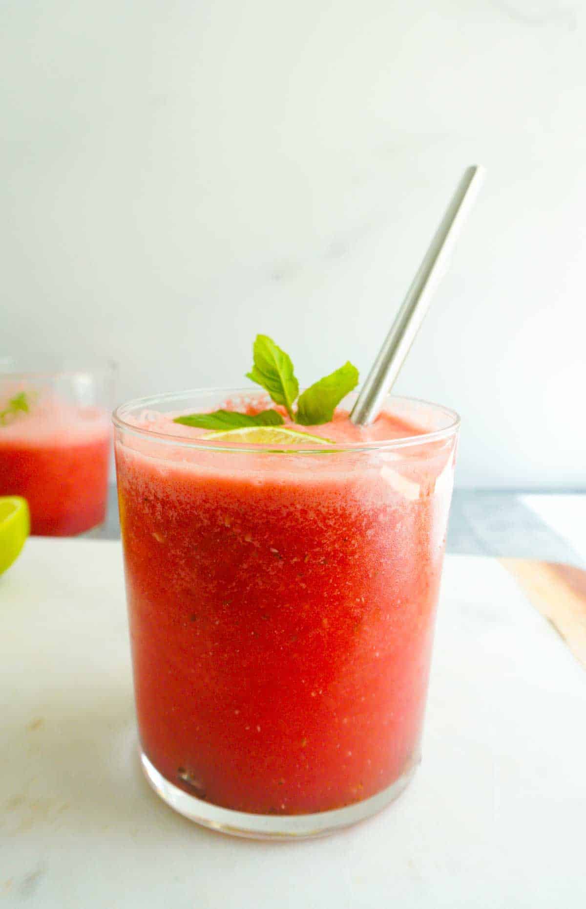 A close up of a glass of watermelon juice. with a slice of lime, some mint and a steel straw. Another glass in the back ground.