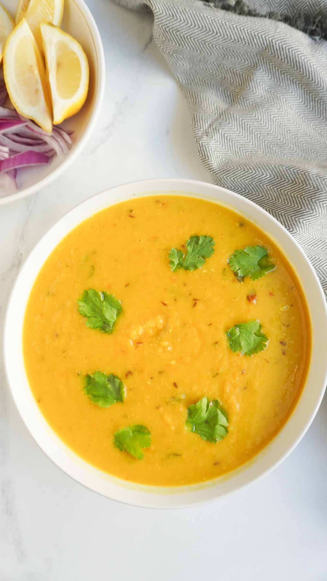 A bowl of tadka masoor daal with cilantro garnish and another bowl with lemon and sliced red onions above it