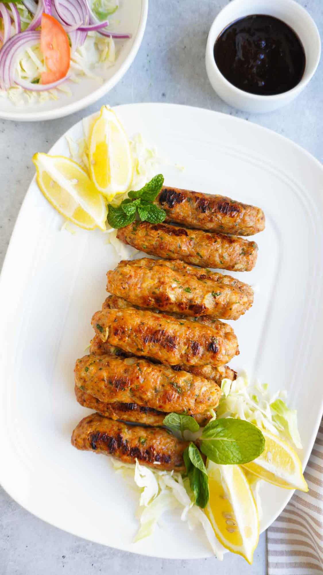 A platter with a row of chicken seekh kebabs, some lemon wedges and mint and a bowl of kachumber and a bowl of tamarind chutney next to it