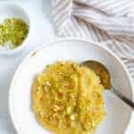 A bowl of almond halwa with a spoon. A small bowl of crushed pistachios to the sde
