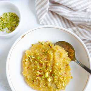 A bowl of almond halwa with a spoon. A small bowl of crushed pistachios to the sde