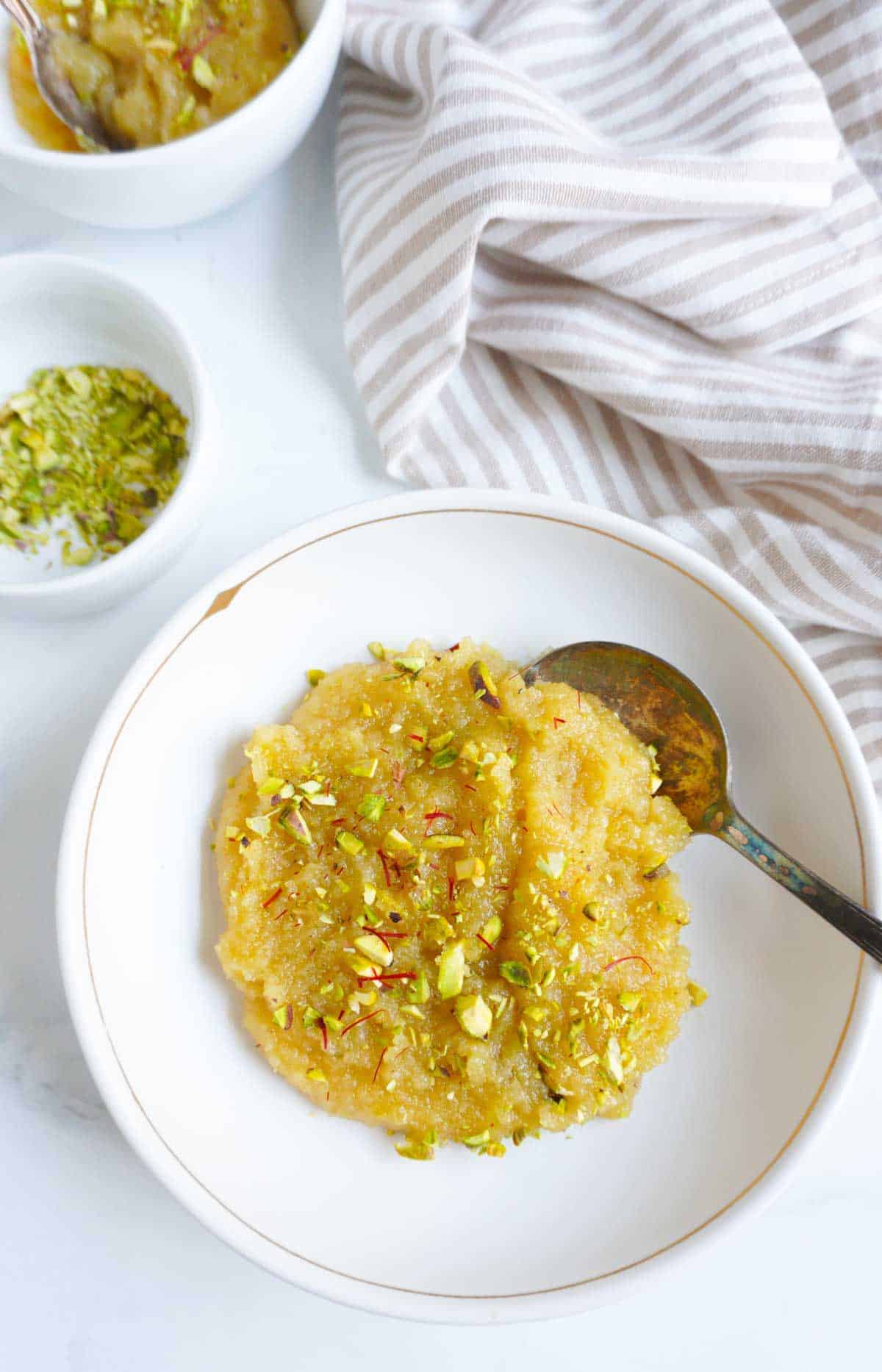A bowl of almond halwa with a spoon. A small bowl of crushed pistachios to the sde