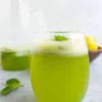 A glass of keto mint lemonade. A bowl of lemons and a jug of lemonade in the back ground.